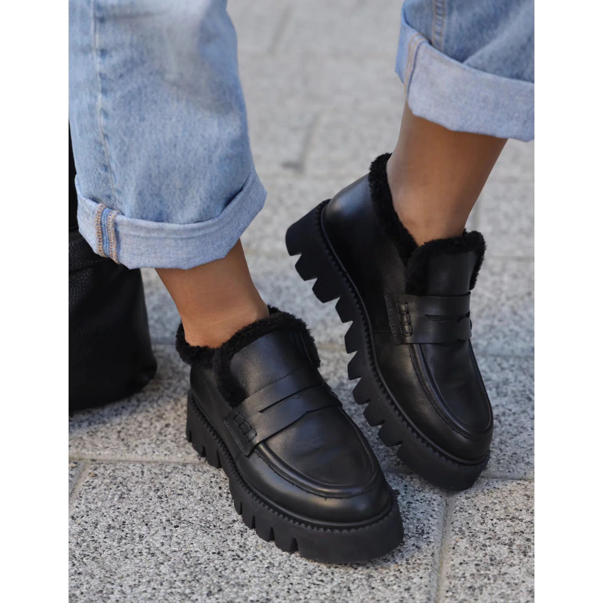 black slip-on loafers with chunky soles and fuzzy lining worn with rolled-up light blue jeans on pavement