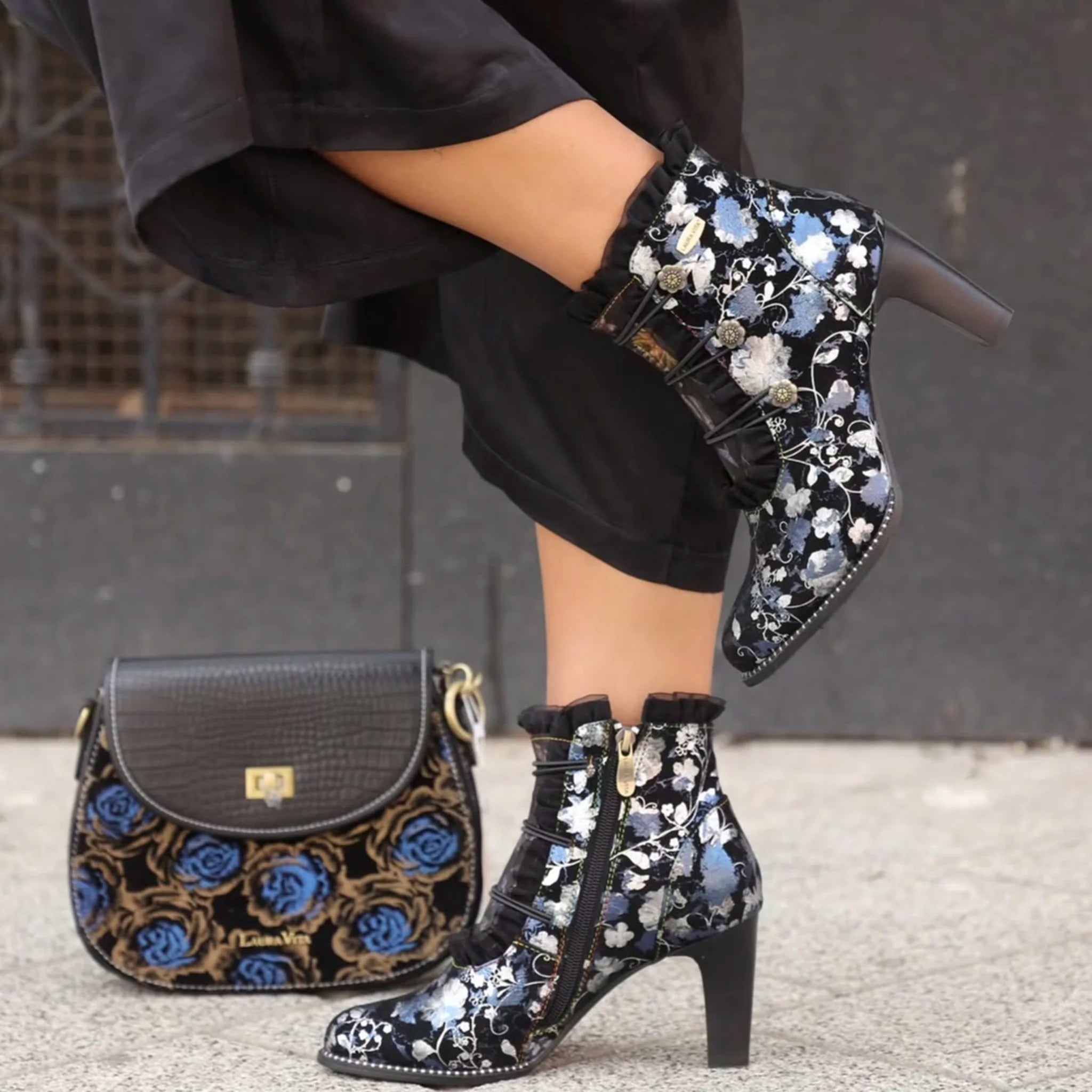 Blue and black floral high-heeled ankle boots paired with a matching floral handbag on a grey sidewalk