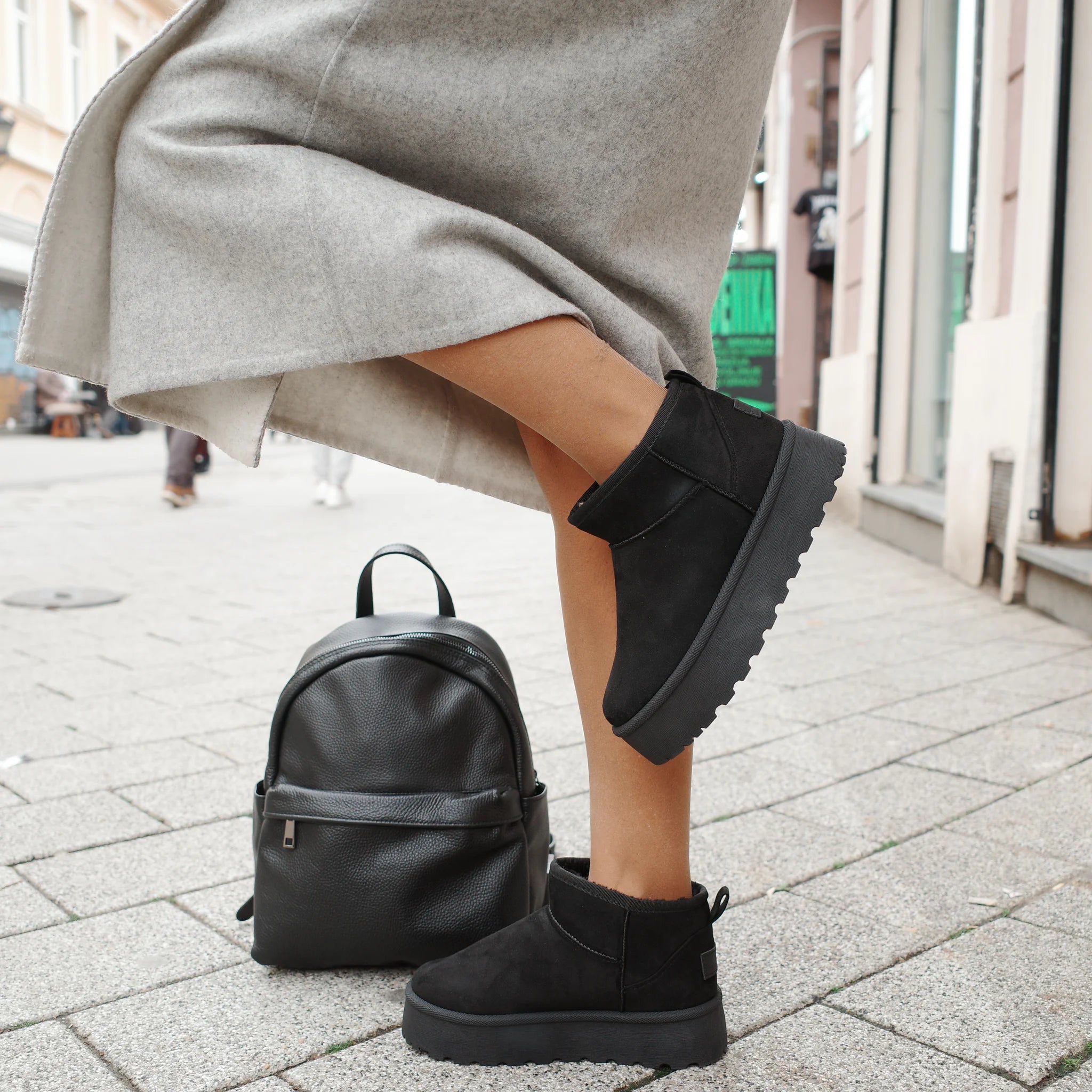 Woman wearing black platform ankle boots and gray coat standing on city sidewalk with black leather backpack