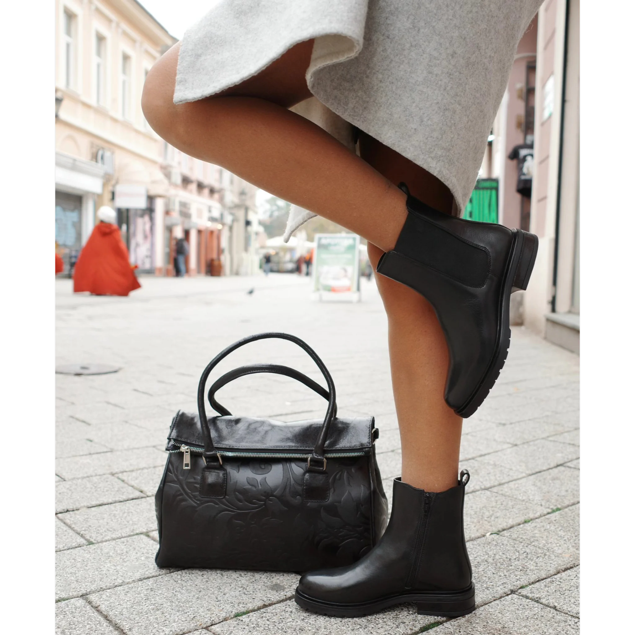 Woman wearing black leather Chelsea boots and gray skirt standing next to black embossed leather handbag on city street