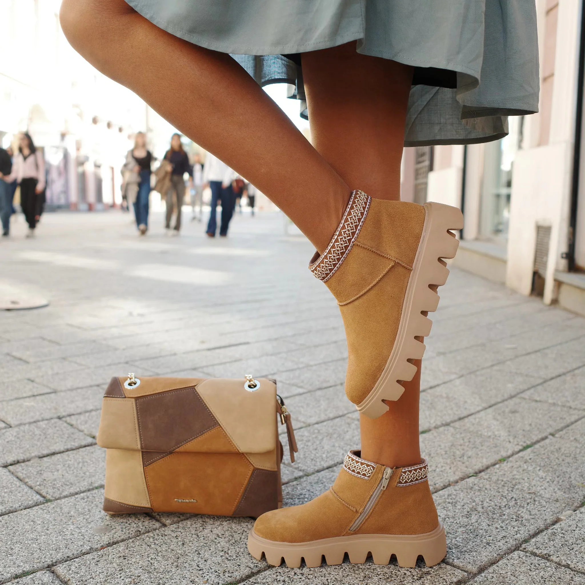Close-up of beige suede ankle boots with decorative trim and thick tread sole, paired with patchwork leather handbag on city street
