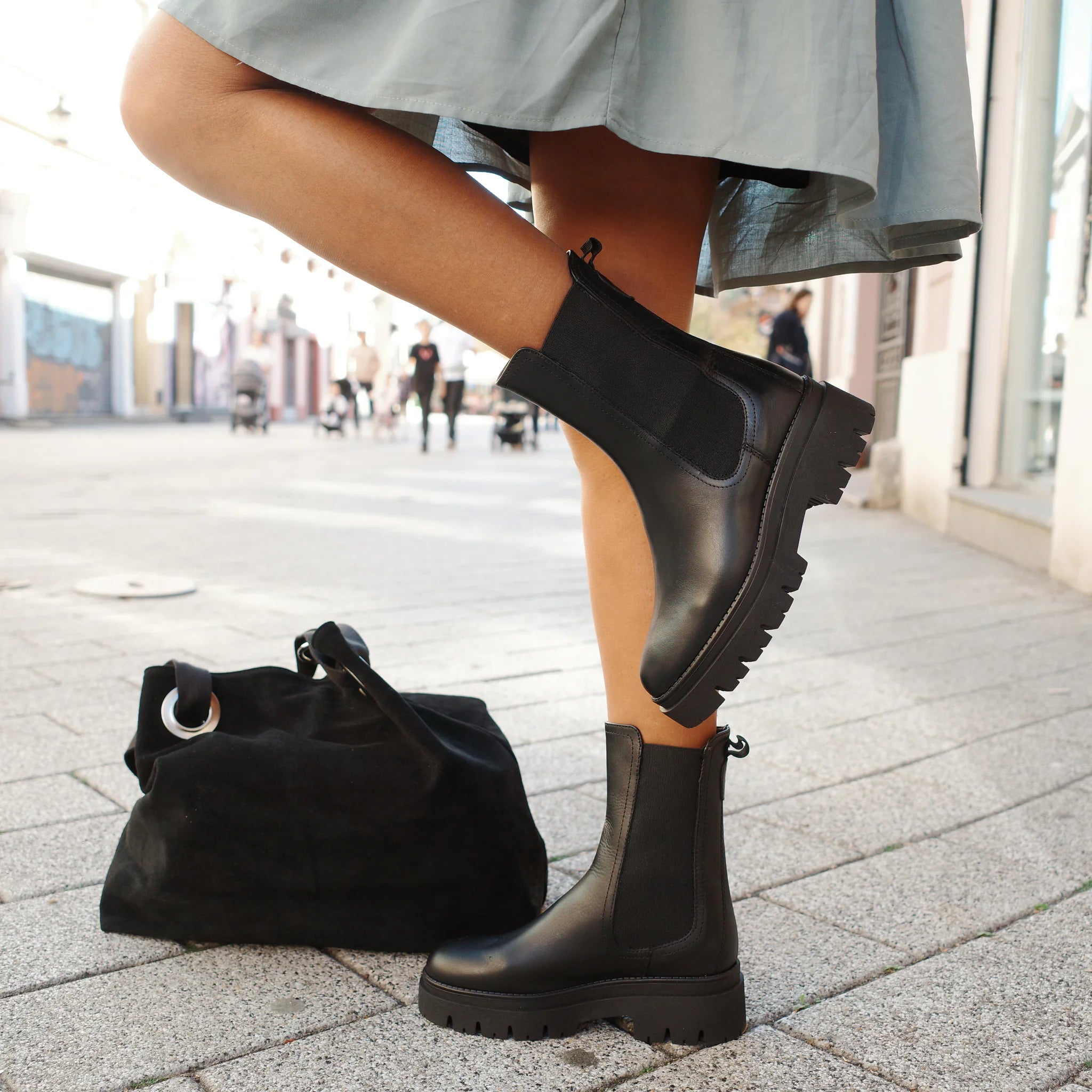 Woman wearing black leather Chelsea boots with chunky soles on city street, next to black suede bag